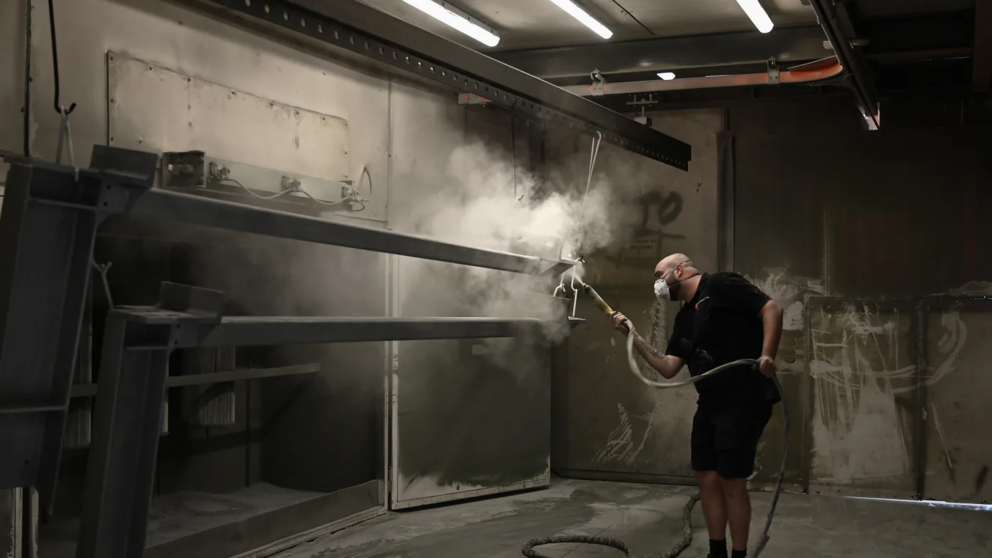 Coater brengt poederlak aan op een stalen ligger, met een wolk poeder in de spuitcabine